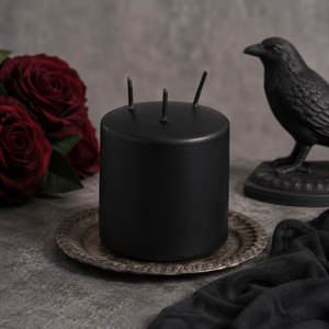 A matte black pillar candle with three wicks sits on a decorative silver plate, styled with red roses and a black raven figurine against a textured gray background.