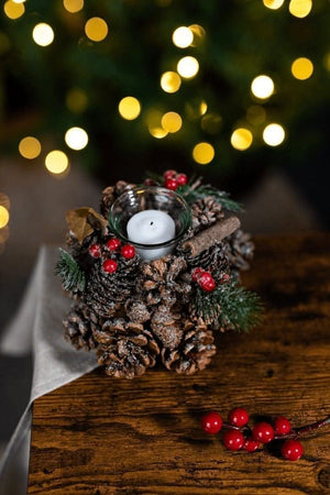 Decorative candle holder with pine cones, berries, and a candle on a wooden surface with blurred lights in the background.