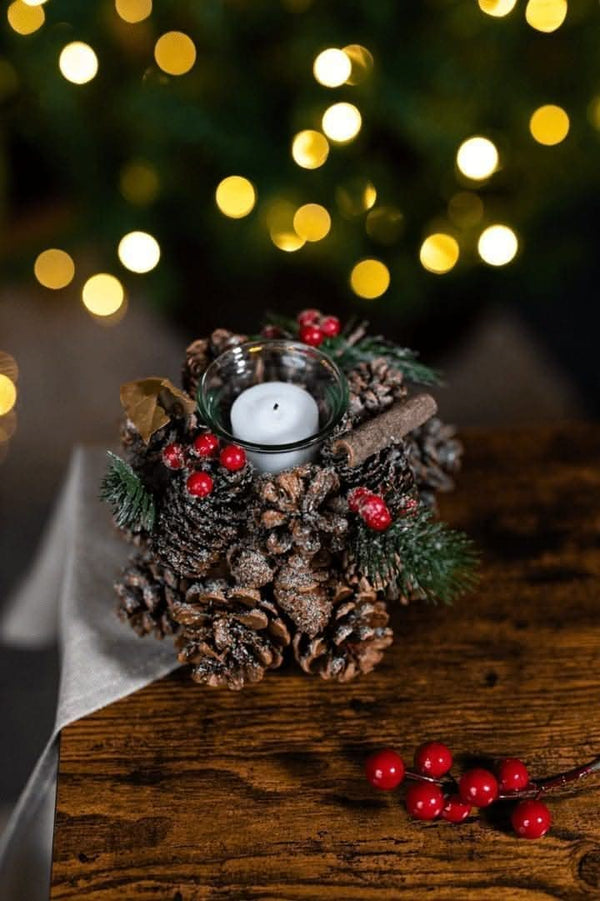 Decorative candle holder with pine cones, berries, and a candle on a wooden surface with blurred lights in the background.