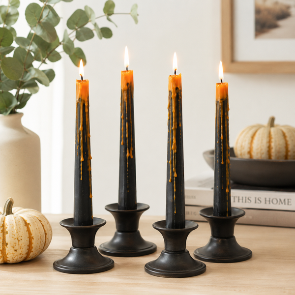 Four black taper candle holders with lit orange taper candles, styled on a wooden table with decorative pumpkins, a vase of eucalyptus, and books in a warm indoor setting.
