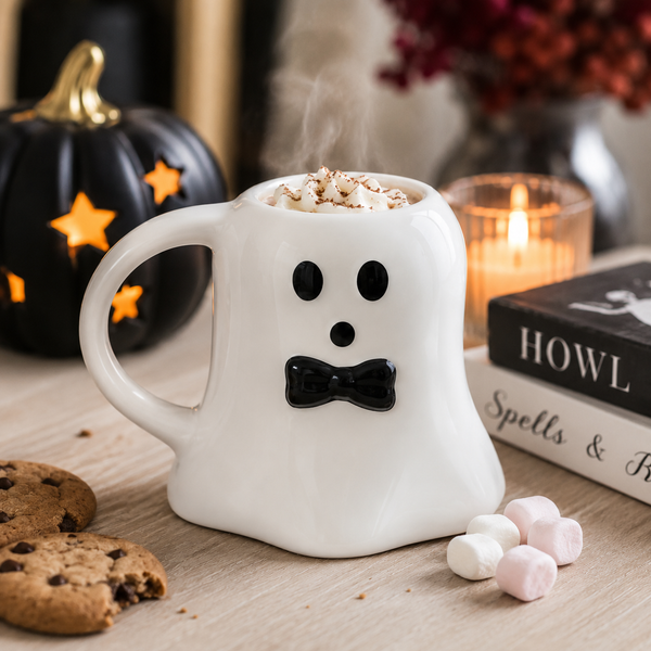 A white ceramic ghost-shaped mug with a black face and bow tie, filled with a hot drink topped with whipped cream and sprinkles, styled on a tabletop with cookies, marshmallows, a black pumpkin, books, and a lit candle.