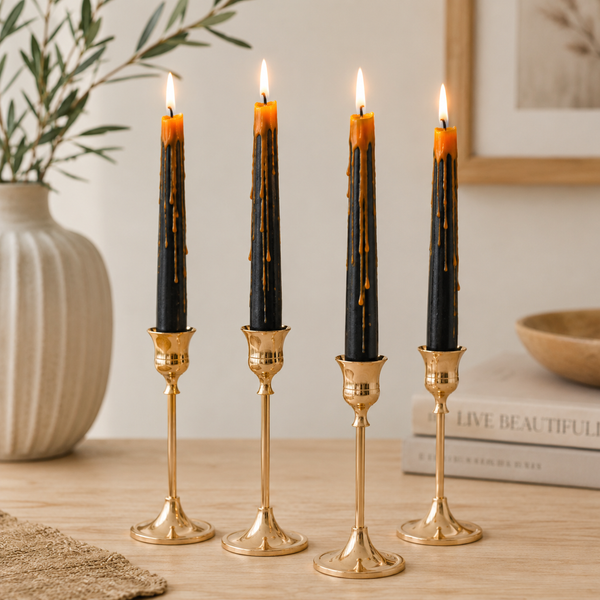 Four black taper candles with orange wax drips in gold candlesticks, arranged on a wooden table in a styled interior setting.