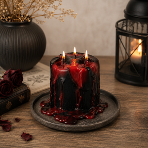 A dark red and black wax candle with dripping wax and three lit wicks, displayed on a rustic wooden table with gothic props including a black lantern, dried flowers, and roses.