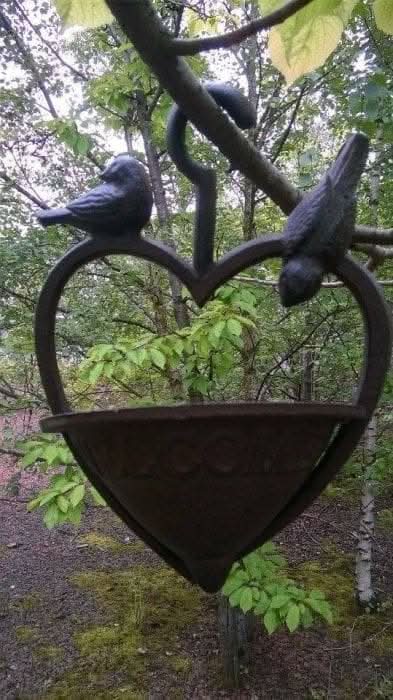A cast iron heart-shaped bird feeder hanging from a tree branch with a bird perched on it.