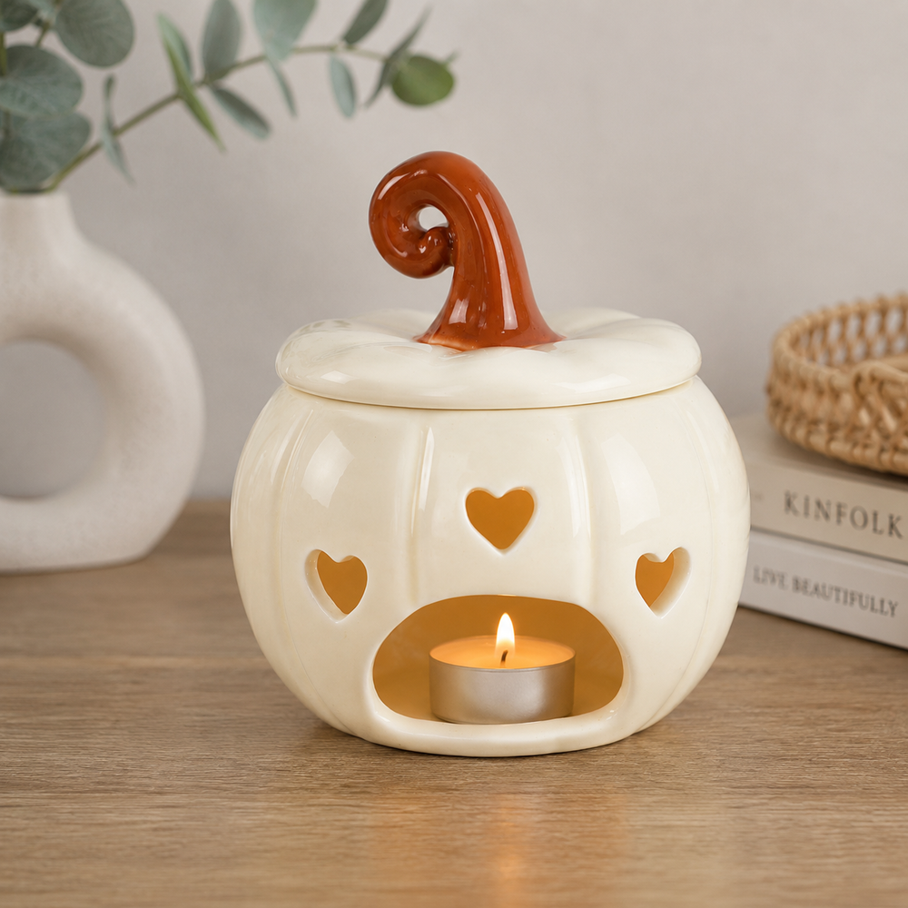 A ceramic pumpkin-shaped candle holder with a lit tea light inside, styled on a wooden surface with a vase and books in the background.