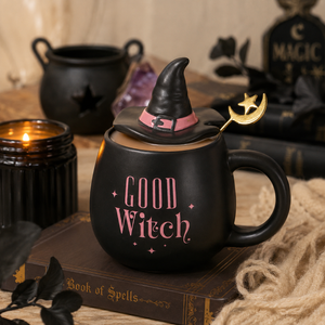 A black ceramic mug shaped like a witch's cauldron with a pointed black hat lid, pink 'GOOD WITCH' text, and a gold crescent moon spoon, styled on a book with candles and witch props in the background.