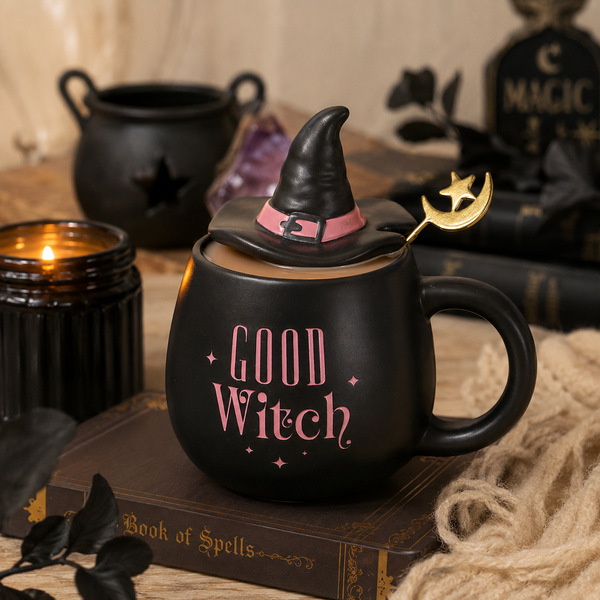 A black ceramic mug shaped like a witch's cauldron with a pointed black hat lid, pink 'GOOD WITCH' text, and a gold crescent moon spoon, styled on a book with candles and witch props in the background.